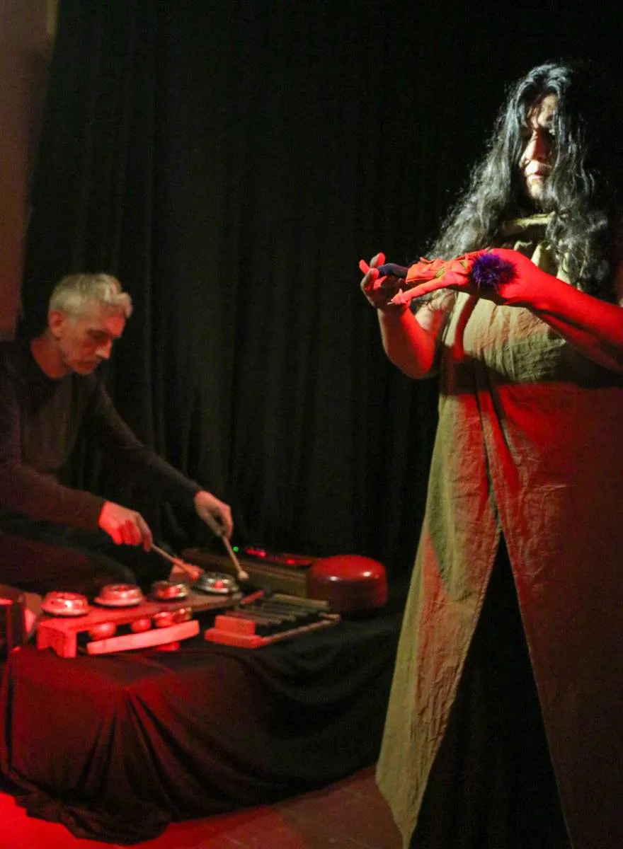 una tura de la escena de la obra emilia en el espacio abierto, adelate un plano americano a la actriz en escena, detrás un musico ejecutando intrumentos atípicos de percusión