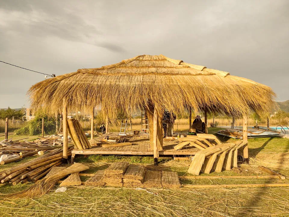 un quincho de paja en construcción, de fondo un paisaje de llanura con vejetación y una pileta